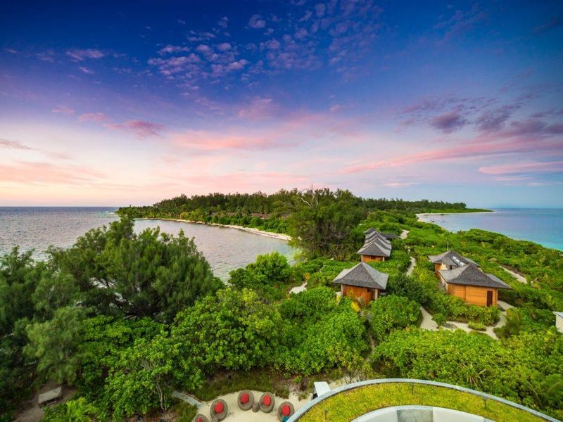 An aerial view of a tropical island resort featuring multiple thatched-roof villas nestled within lush greenery along the coastline.