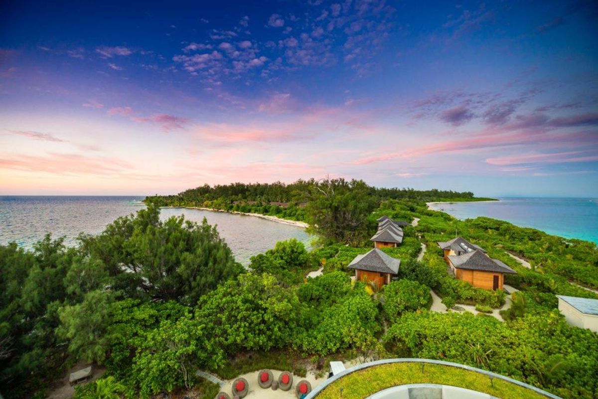 An aerial view of a tropical island resort featuring multiple thatched-roof villas nestled within lush greenery along the coastline.