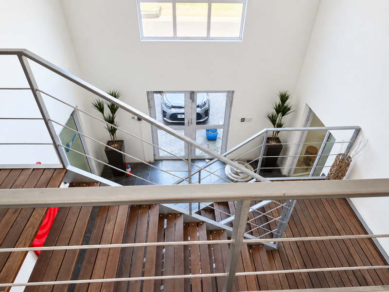 A bright, double-volume entrance foyer featuring a modern timber staircase with metal balustrades and large glass double doors leading to the exterior.