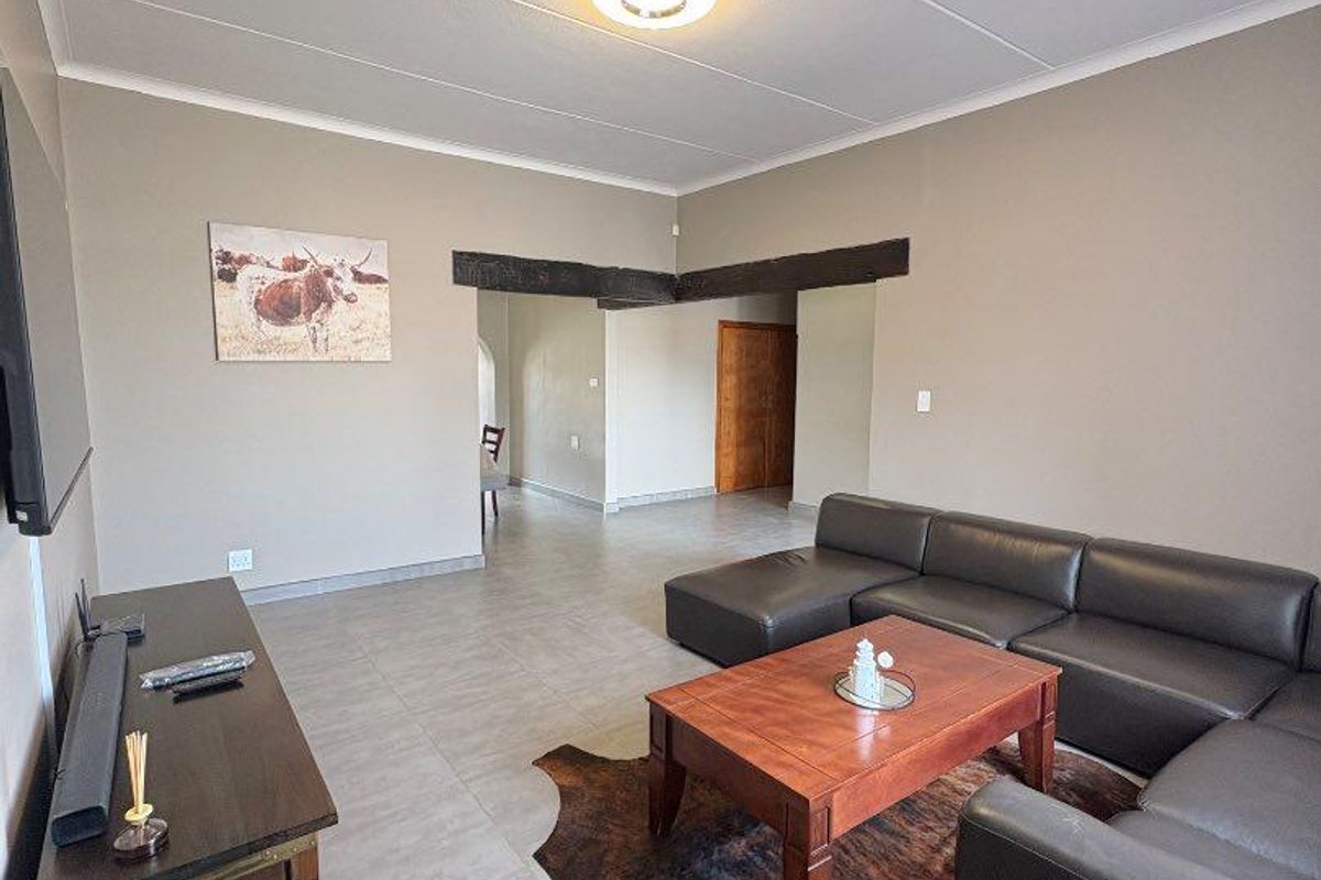 Spacious living room featuring neutral tiled flooring, exposed timber ceiling beams, and a clean, open-plan layout leading to adjacent areas.