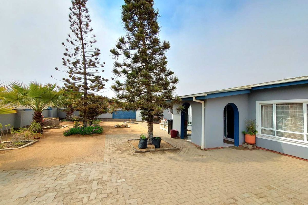 A rear exterior view of the property featuring a paved courtyard, mature trees, and a covered entrance with arched architectural detailing.