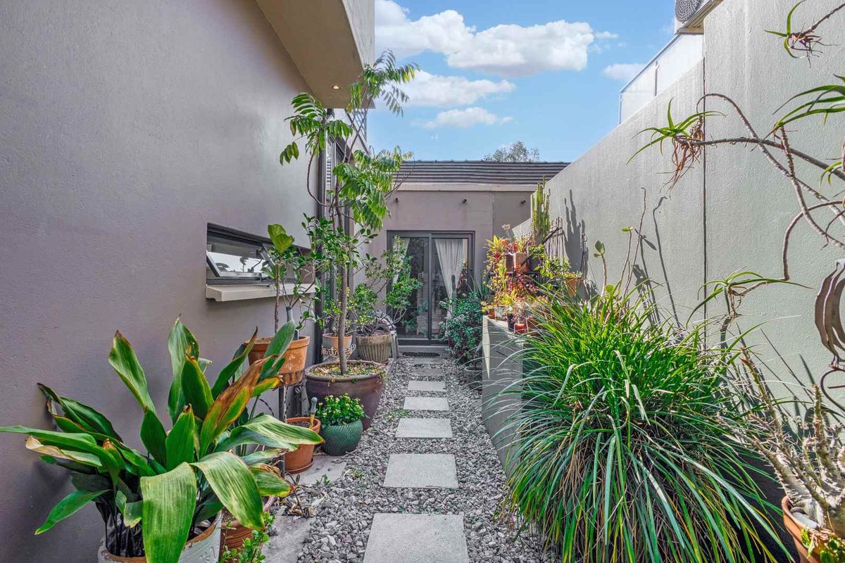 A narrow rear courtyard featuring a stone paver walkway set in gravel, bordered by retaining walls and providing access to the home via sliding doors.