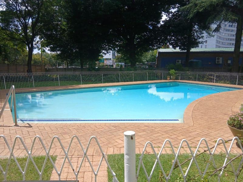 A well-maintained outdoor swimming pool area featuring paved surrounds and secure perimeter fencing, set within a landscaped garden environment.