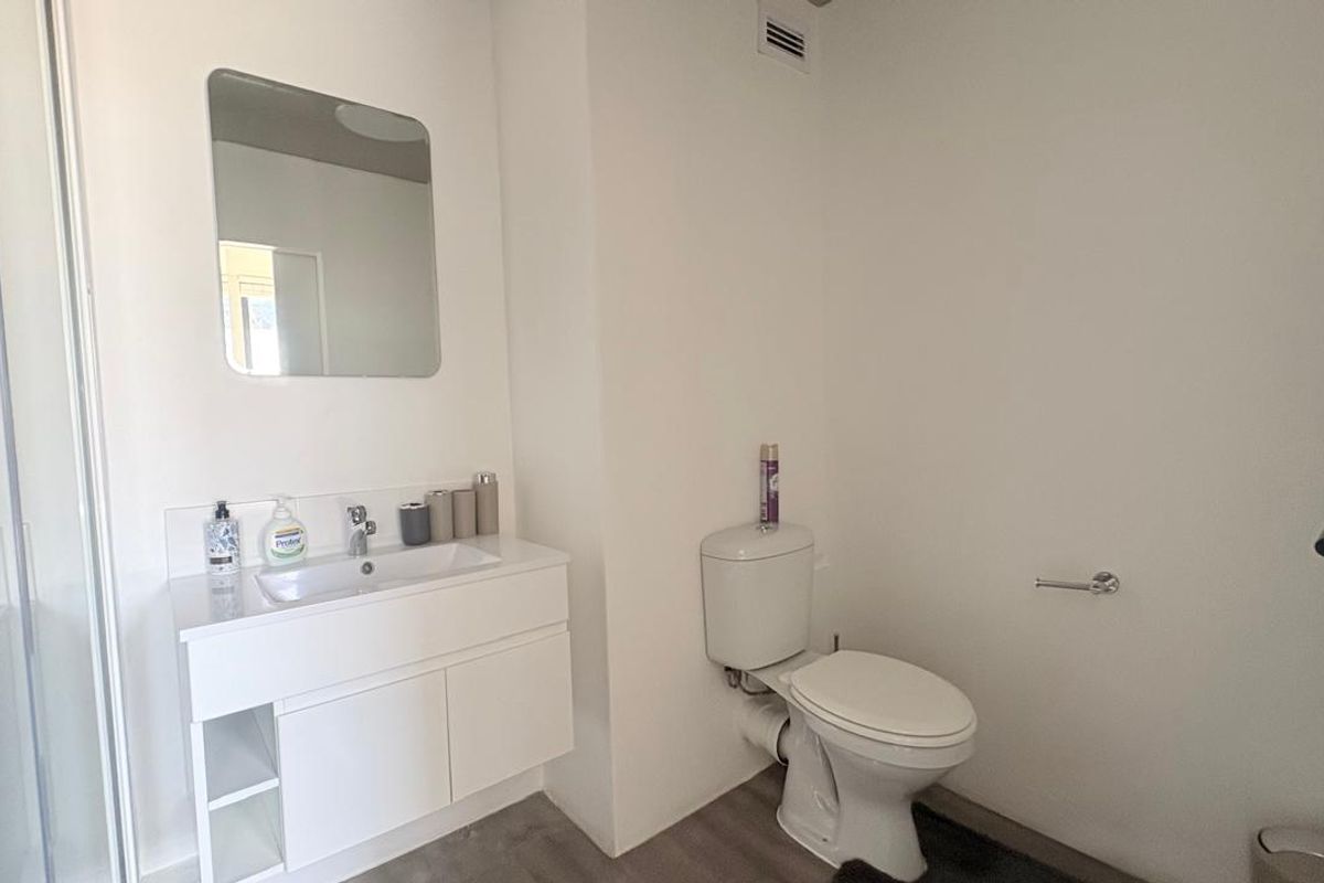 A clean, modern bathroom featuring a white vanity with integrated basin, wall-mounted mirror, toilet, and a glass-enclosed shower area.