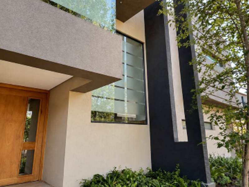 Modern architectural front facade featuring a timber entrance door, glass balcony balustrade, and clean geometric lines with integrated garden landscaping.