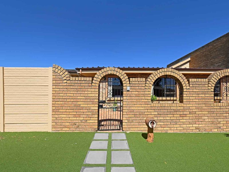 Front exterior view featuring a face brick facade with arched window openings, a secure metal gate, paved walkway, and well-maintained artificial lawn.