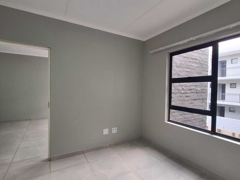 A clean, empty bedroom featuring neutral grey walls, large aluminium-framed windows, and modern tiled flooring, offering a bright and versatile living space.