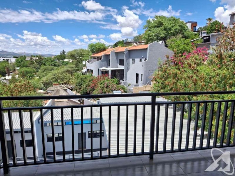 A spacious balcony featuring a metal balustrade and tiled flooring, offering an elevated view of the surrounding residential complex and landscape.