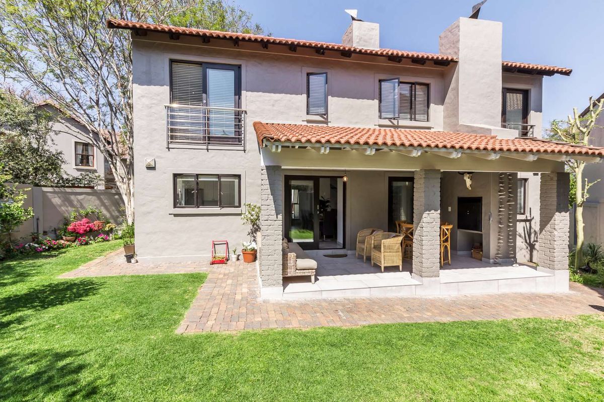 Rear view of a two-story house featuring a covered patio with a built-in braai, tiled flooring, and a landscaped garden.