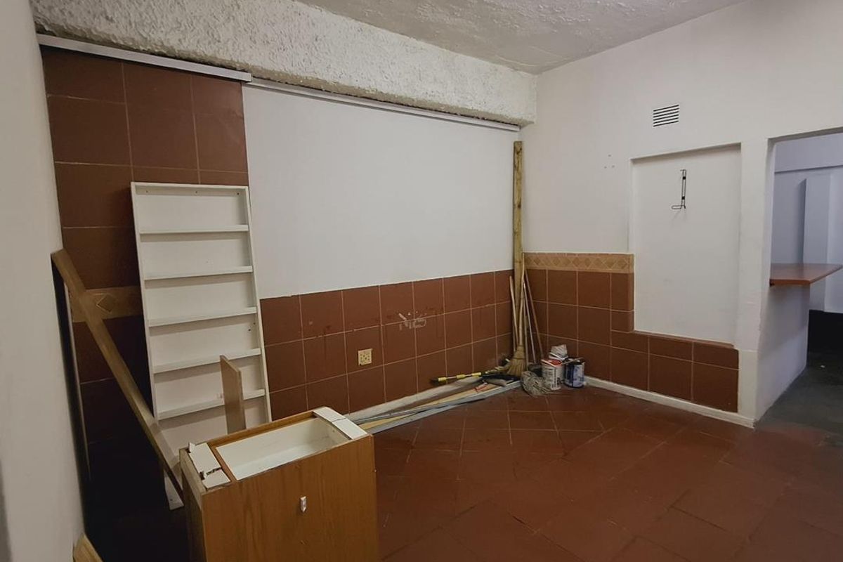 An empty, dated kitchen area featuring brown tiled flooring and wall splashbacks, with a pass-through window connecting to an adjacent space.