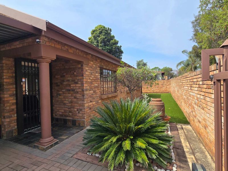A well-maintained rear exterior featuring a face brick facade, covered patio area, paved walkway, and a neat artificial lawn enclosed by brick boundary walls.