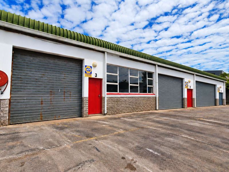 Industrial warehouse exterior featuring multiple roller shutter doors, red access doors, and a concrete forecourt area with security signage.