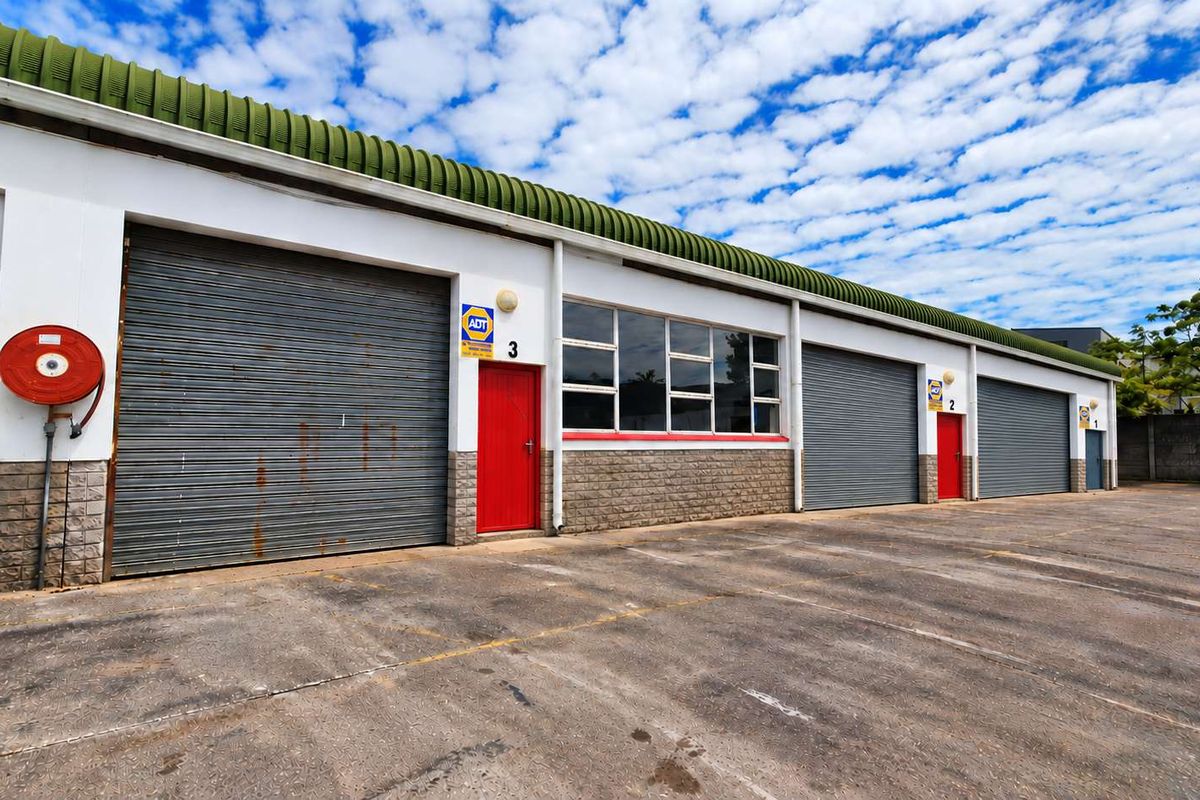 Industrial warehouse exterior featuring multiple roller shutter doors, red access doors, and a concrete forecourt area with security signage.