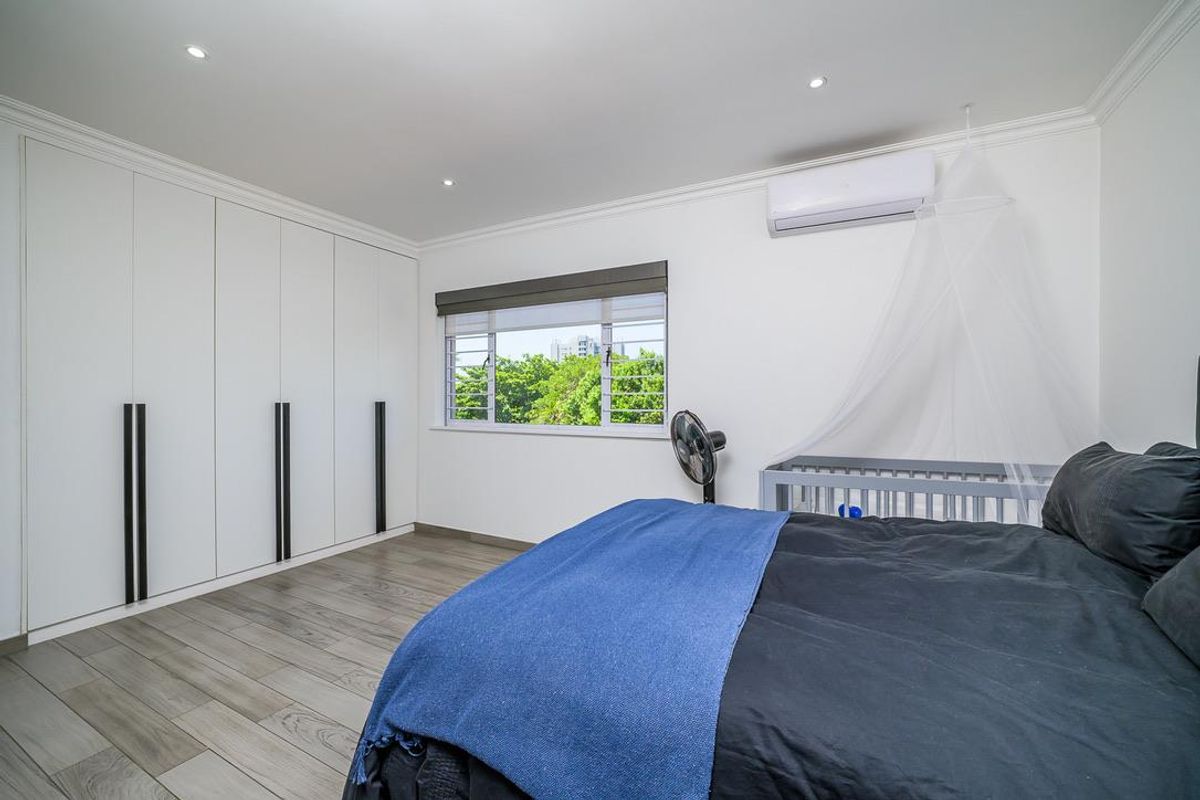 A bright bedroom featuring modern wood-look flooring, extensive built-in floor-to-ceiling cabinetry, recessed downlighting, and a wall-mounted air conditioning unit.