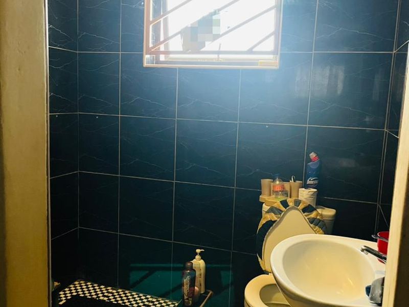 A compact bathroom featuring dark tiled walls, a tiled shower area with a checkered floor, a pedestal basin, and a toilet.