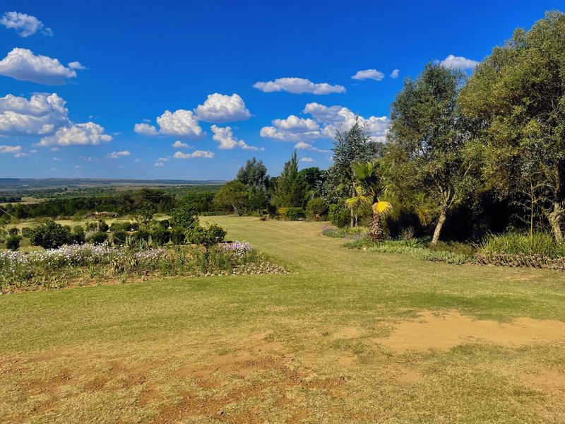 A well-maintained expansive garden featuring mature trees, landscaped flower beds, and scenic open views across the small holding landscape.