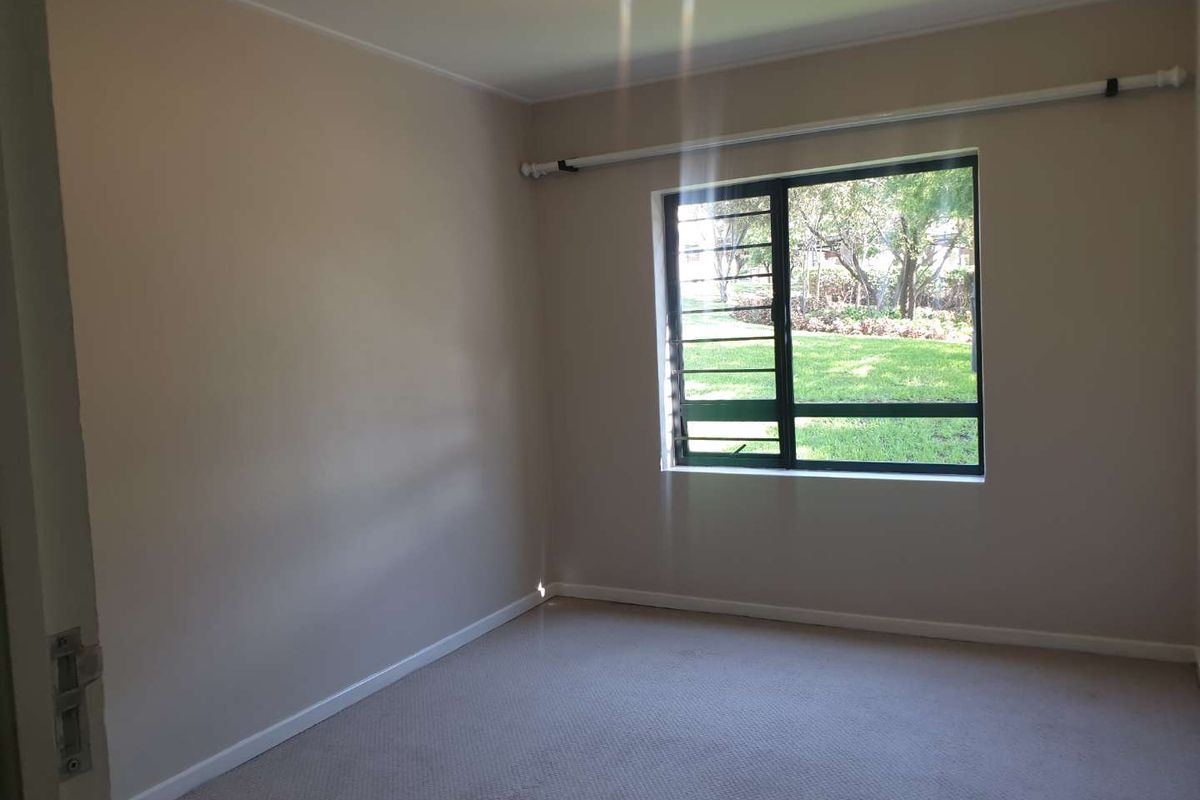 A clean, empty bedroom featuring neutral walls, carpeted flooring, and a window with security bars overlooking a garden area.