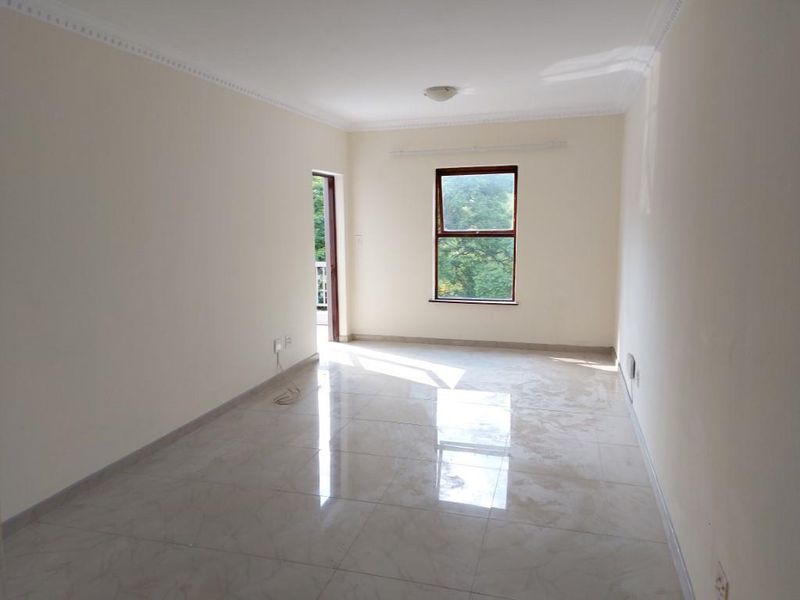 A bright, empty living room featuring neutral tiled flooring, cornice detailing, and natural light from a window and adjacent doorway.