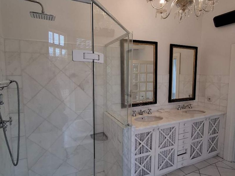 A well-appointed bathroom featuring a double vanity with marble countertops, a glass-enclosed shower with rainfall fixture, and elegant chandelier lighting.