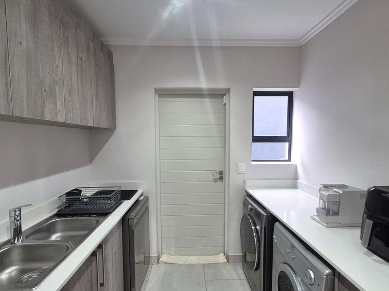 A modern, well-maintained scullery featuring sleek wood-finish cabinetry, stone countertops, a double basin sink, and dedicated plumbing connections for laundry appliances.