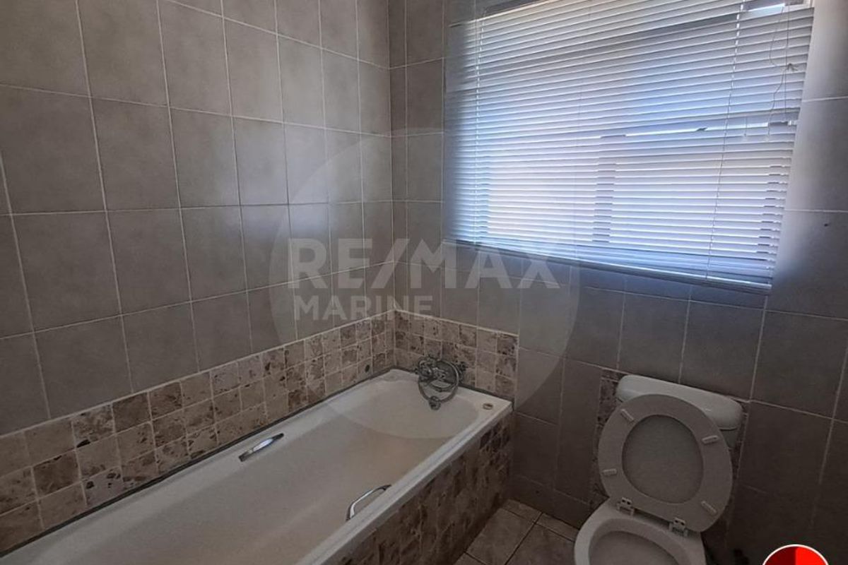 A functional bathroom featuring a built-in bathtub with tiled surround, a standard toilet, and a window with fitted blinds.