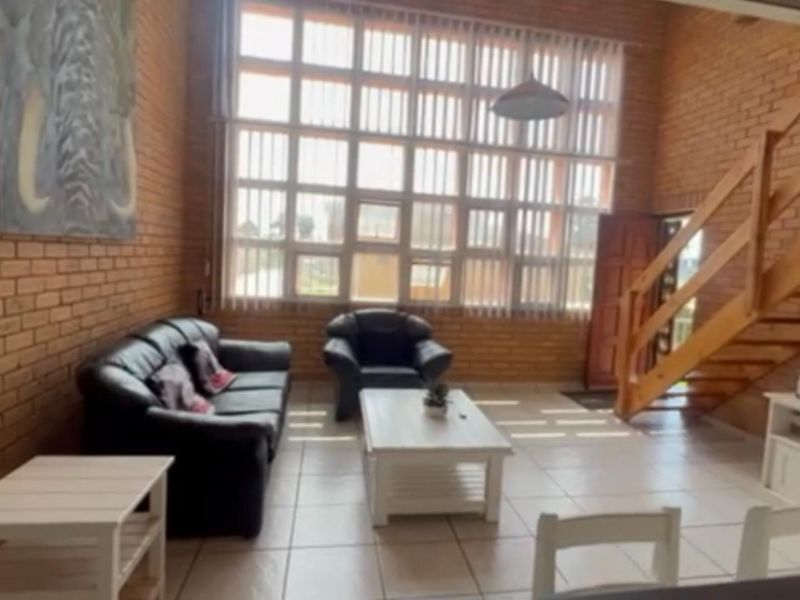Spacious living area featuring double-volume exposed brick walls, large windows with vertical blinds, and a wooden staircase leading to the upper level.