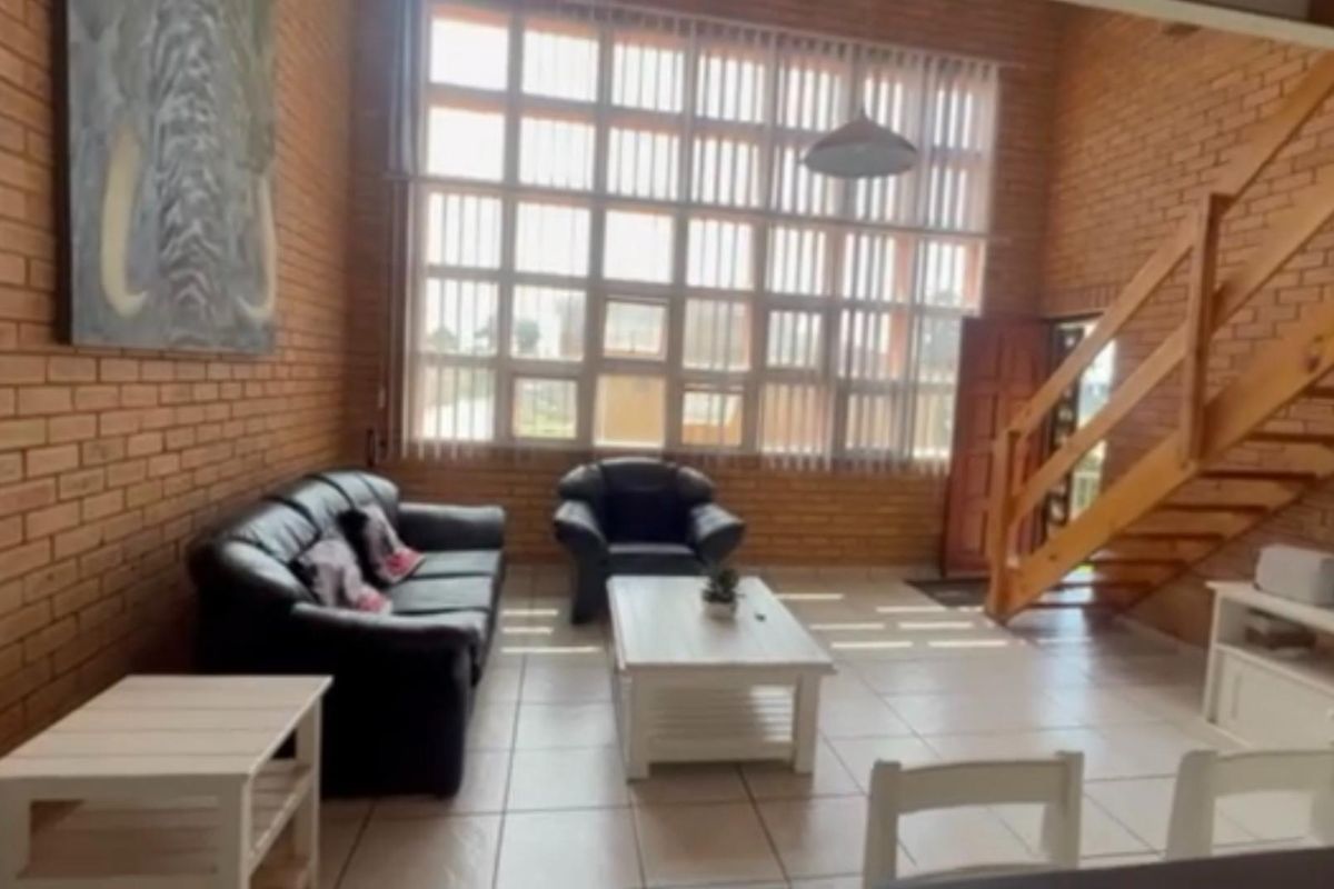 Spacious living area featuring double-volume exposed brick walls, large windows with vertical blinds, and a wooden staircase leading to the upper level.