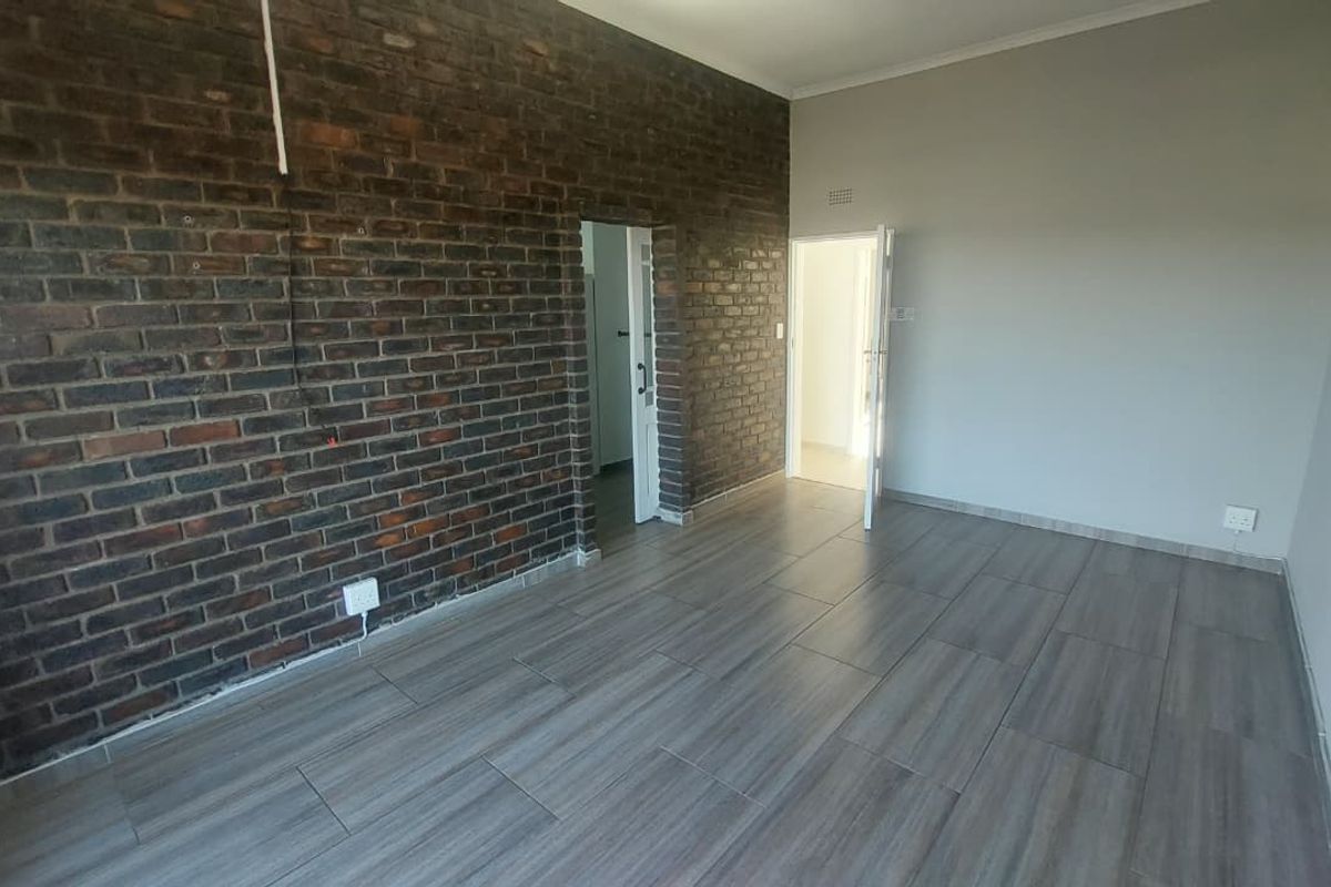 An empty living area featuring a prominent exposed brick feature wall, modern wood-look floor tiles, and access to adjacent rooms.