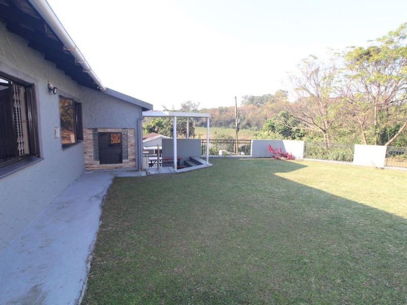 Rear exterior view featuring a spacious lawn, a covered patio area, and a built-in braai, overlooking a natural landscape.