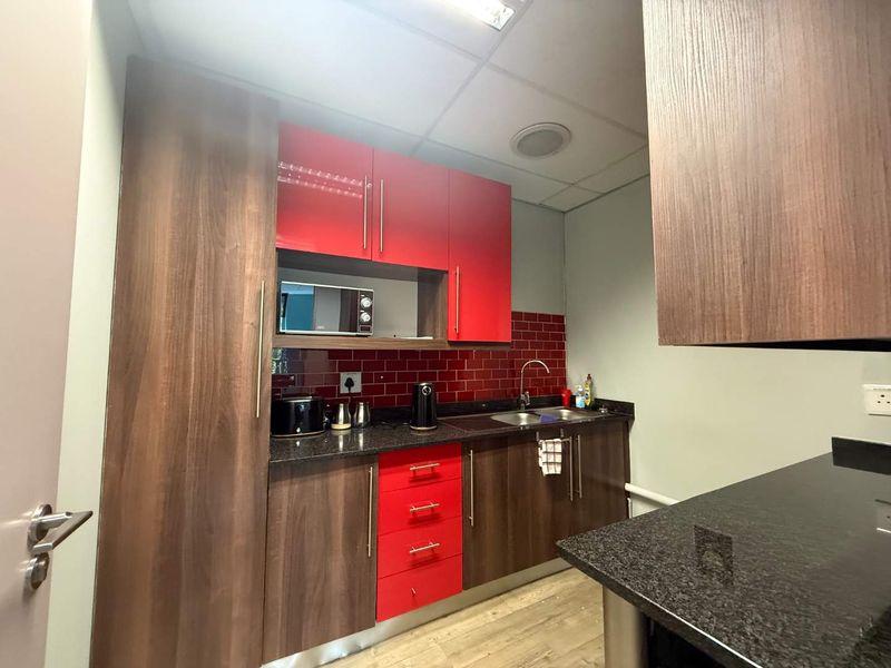 A compact office kitchenette featuring modern wood-grain and red cabinetry, granite countertops, a stainless steel sink, and a vibrant red tiled backsplash.