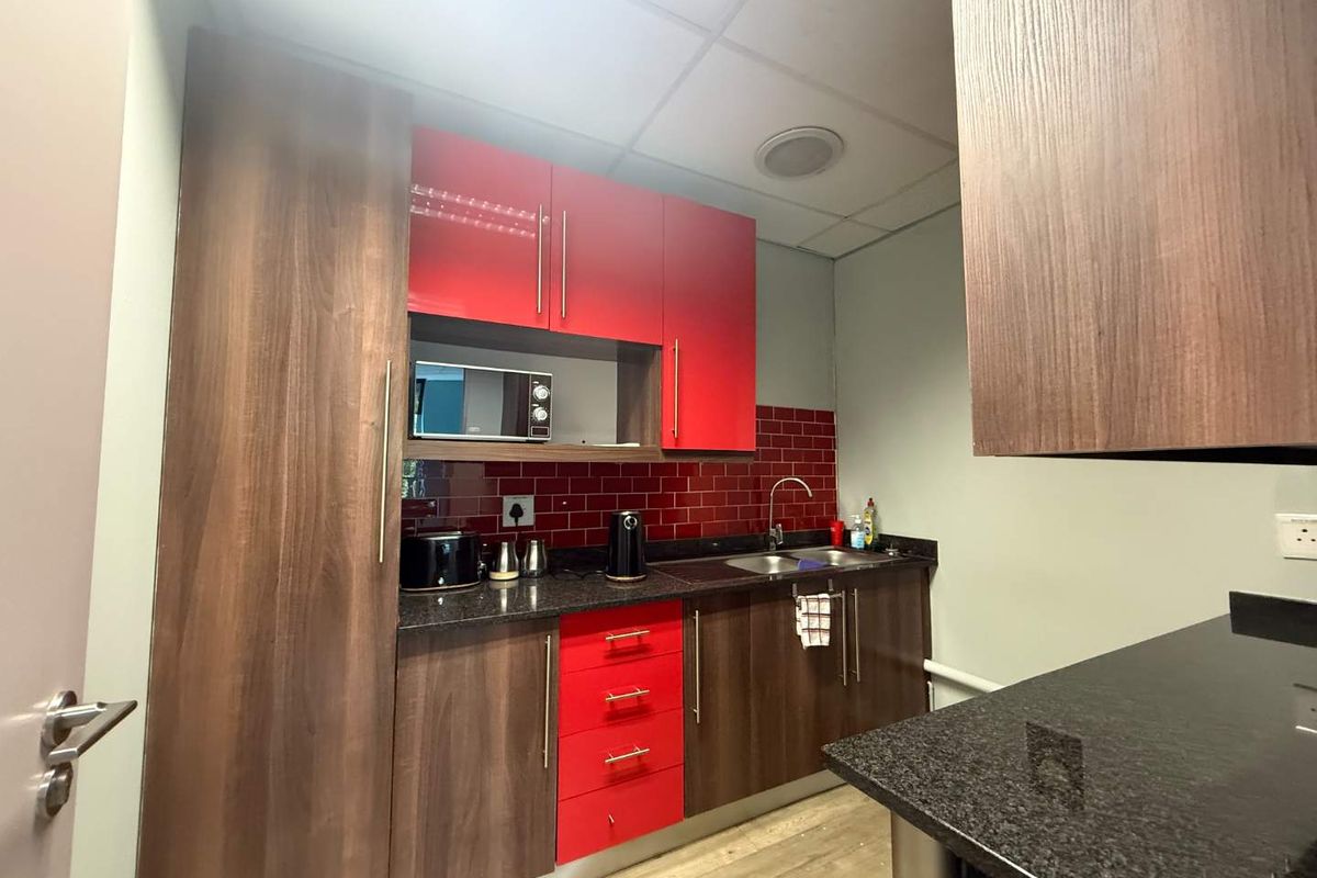 A compact office kitchenette featuring modern wood-grain and red cabinetry, granite countertops, a stainless steel sink, and a vibrant red tiled backsplash.