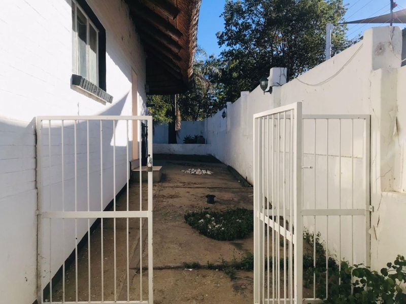A narrow rear courtyard area featuring a thatched roof overhang, concrete paving, and a white perimeter wall with security gate access.