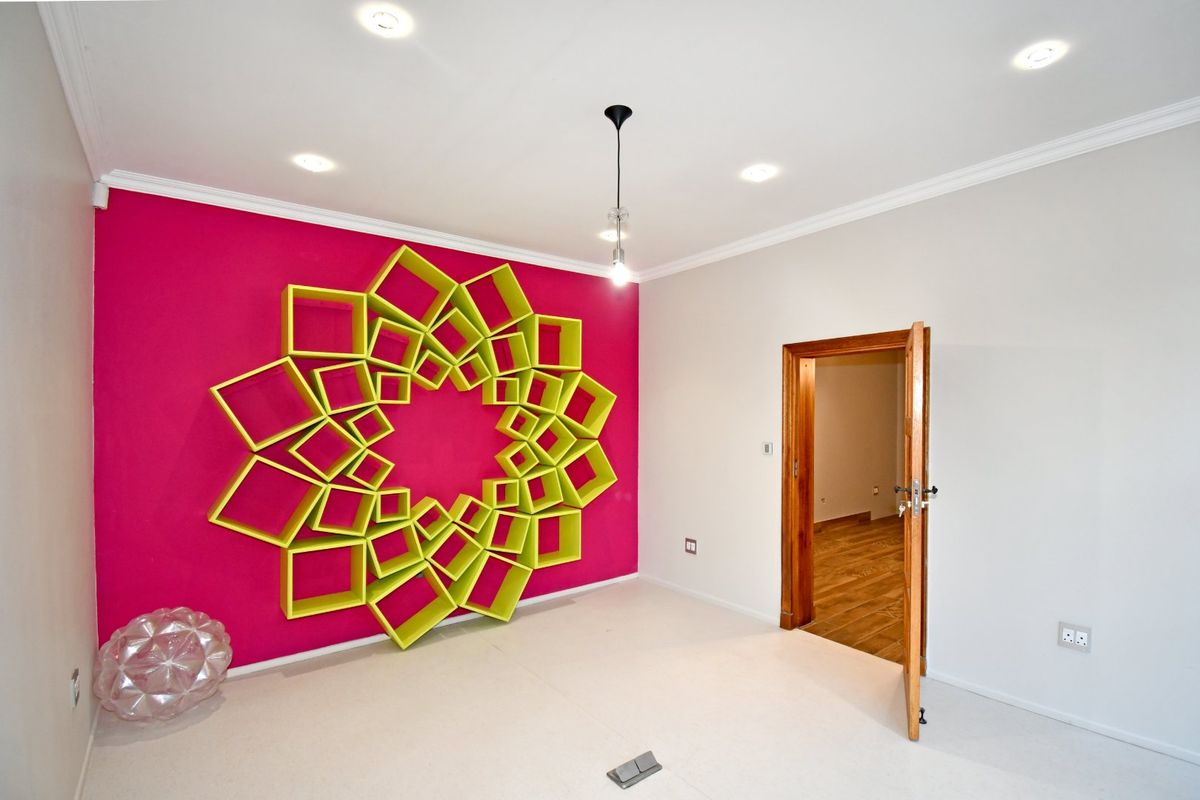A bright, empty bedroom featuring a striking geometric wall installation, recessed ceiling lighting, and a solid timber door frame.