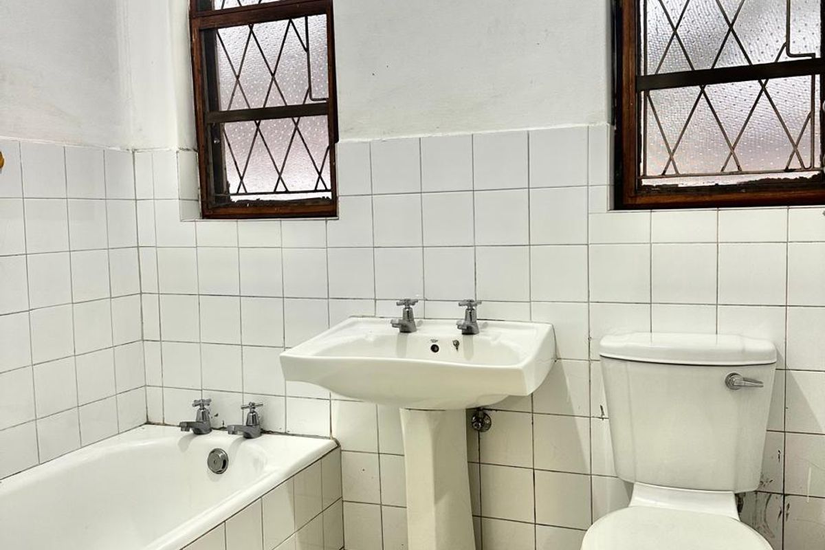 A dated bathroom featuring a pedestal basin, built-in bathtub, and toilet, with original leaded glass windows and white ceramic tiling throughout.