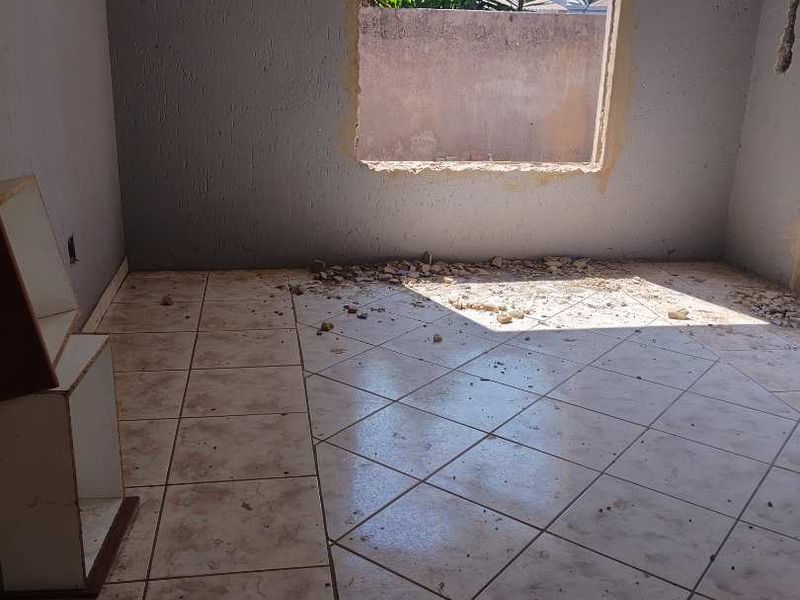 An empty bedroom requiring renovation, featuring tiled flooring, decorative cornice, and a window opening with visible structural damage to the ceiling and wall.