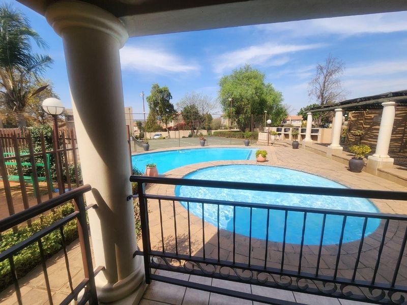 Outdoor pool area featuring a paved patio, built-in braai facilities, and metal balustrades, overlooking a communal swimming pool and tennis court.