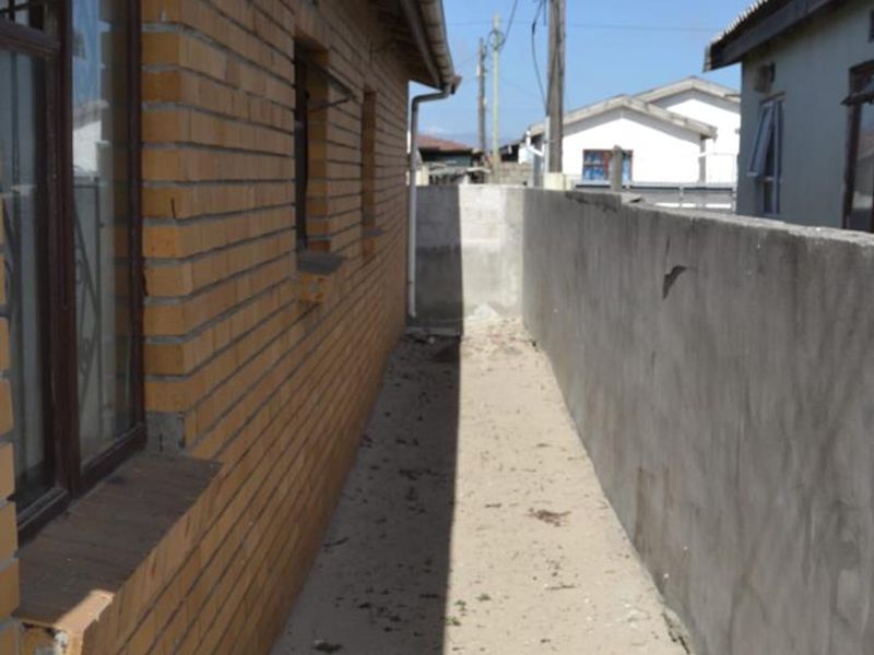 A narrow side passage between the brick house exterior and a concrete boundary wall, showing basic exterior finishes.