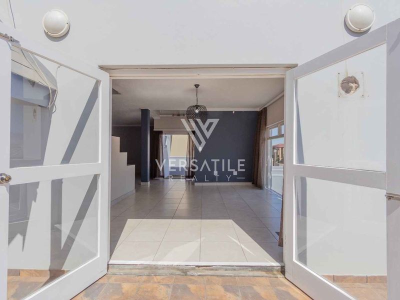 An open-plan living area viewed from an outdoor patio through double glass doors, featuring tiled flooring and a staircase.