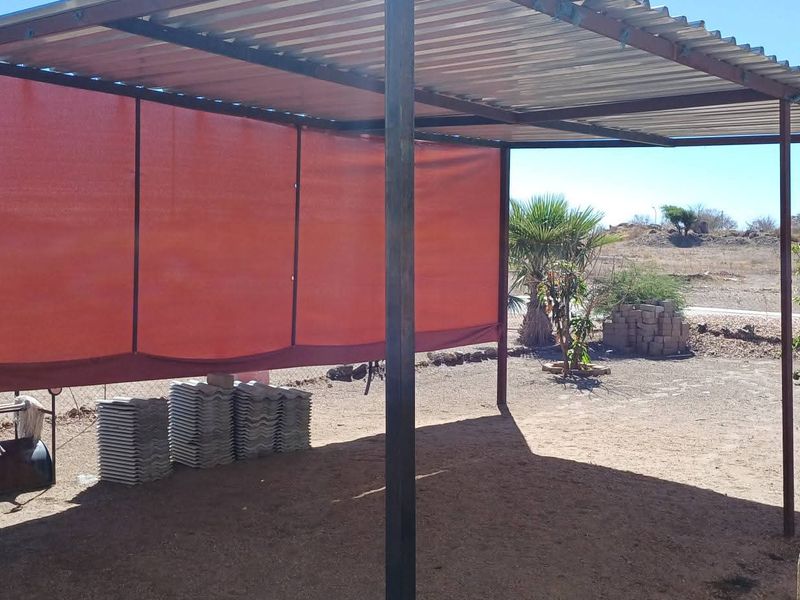 A simple outdoor covered patio area featuring a corrugated metal roof supported by metal posts and a privacy shade screen.