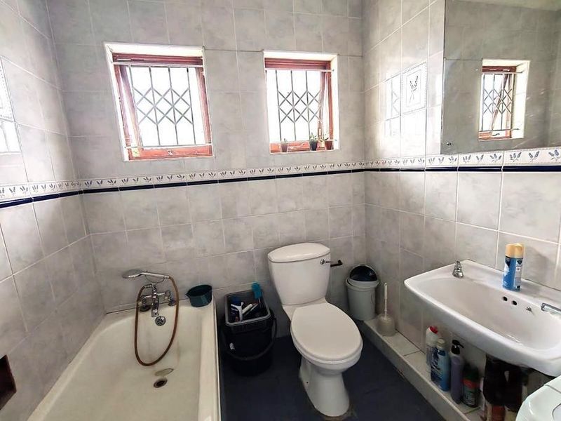 A functional bathroom featuring a bathtub, pedestal basin, and toilet, finished with tiled walls and security bars on the windows.
