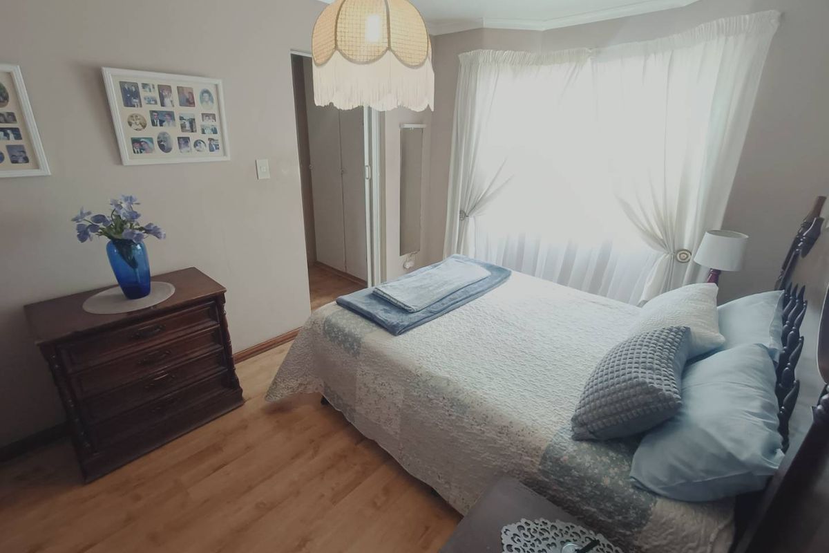 A cozy bedroom featuring light wood-look flooring, neutral wall tones, and a large window with sheer curtains, providing a bright and comfortable living space.