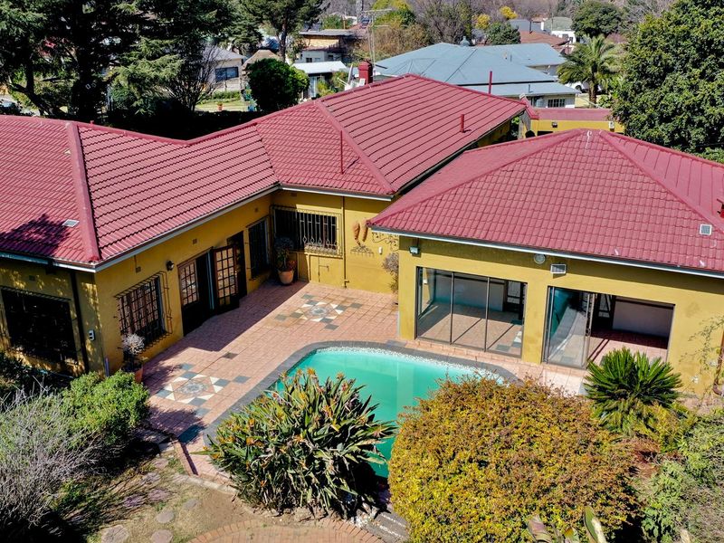 Aerial view of the property featuring a swimming pool, paved patio area, and a red metal roof structure surrounded by established garden landscaping.