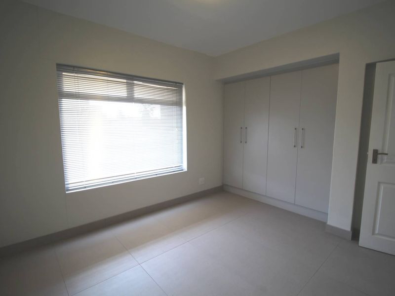 A bright, empty bedroom featuring neutral tiled flooring, built-in cupboards, and venetian blinds, offering a clean and versatile space.