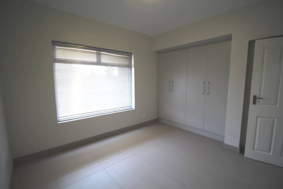 A bright, empty bedroom featuring neutral tiled flooring, built-in cupboards, and venetian blinds, offering a clean and versatile space.