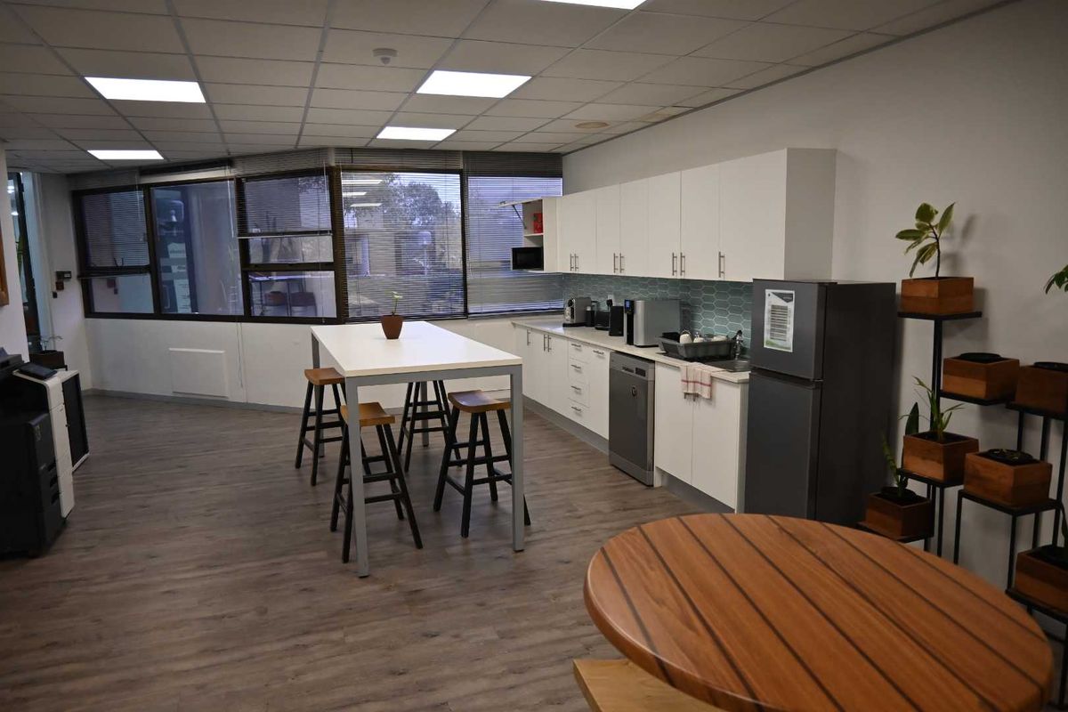 A functional office kitchenette featuring white cabinetry, a tiled backsplash, laminate flooring, and a central high-top table for casual dining or meetings.