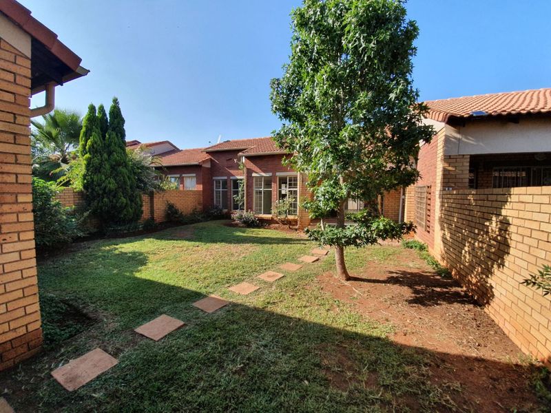 A private rear garden featuring a brick boundary wall, established trees, and a paved walkway leading towards the townhouse structure with a tiled roof.