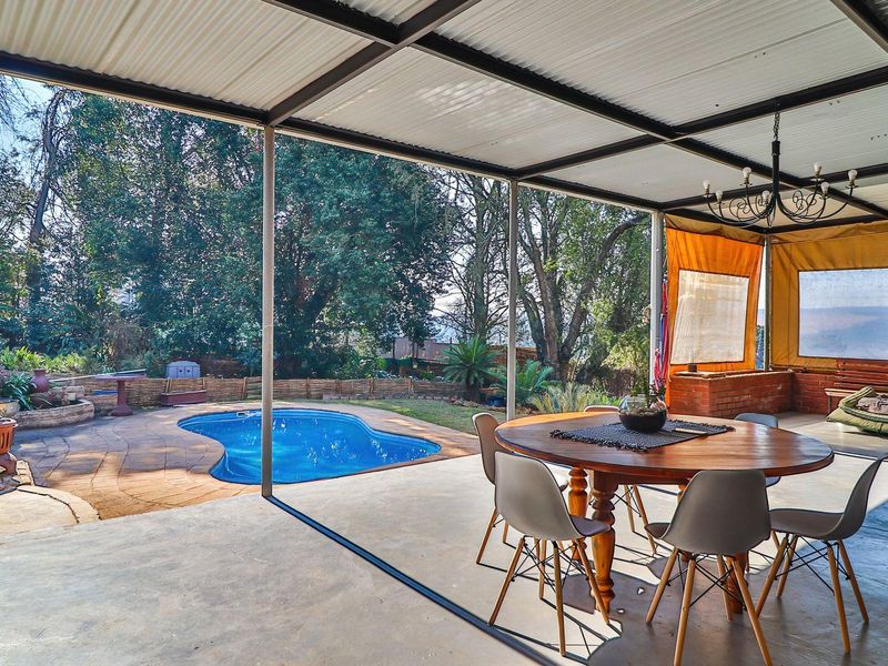 A covered outdoor patio area featuring concrete flooring and a corrugated roof, overlooking a private swimming pool and landscaped garden.