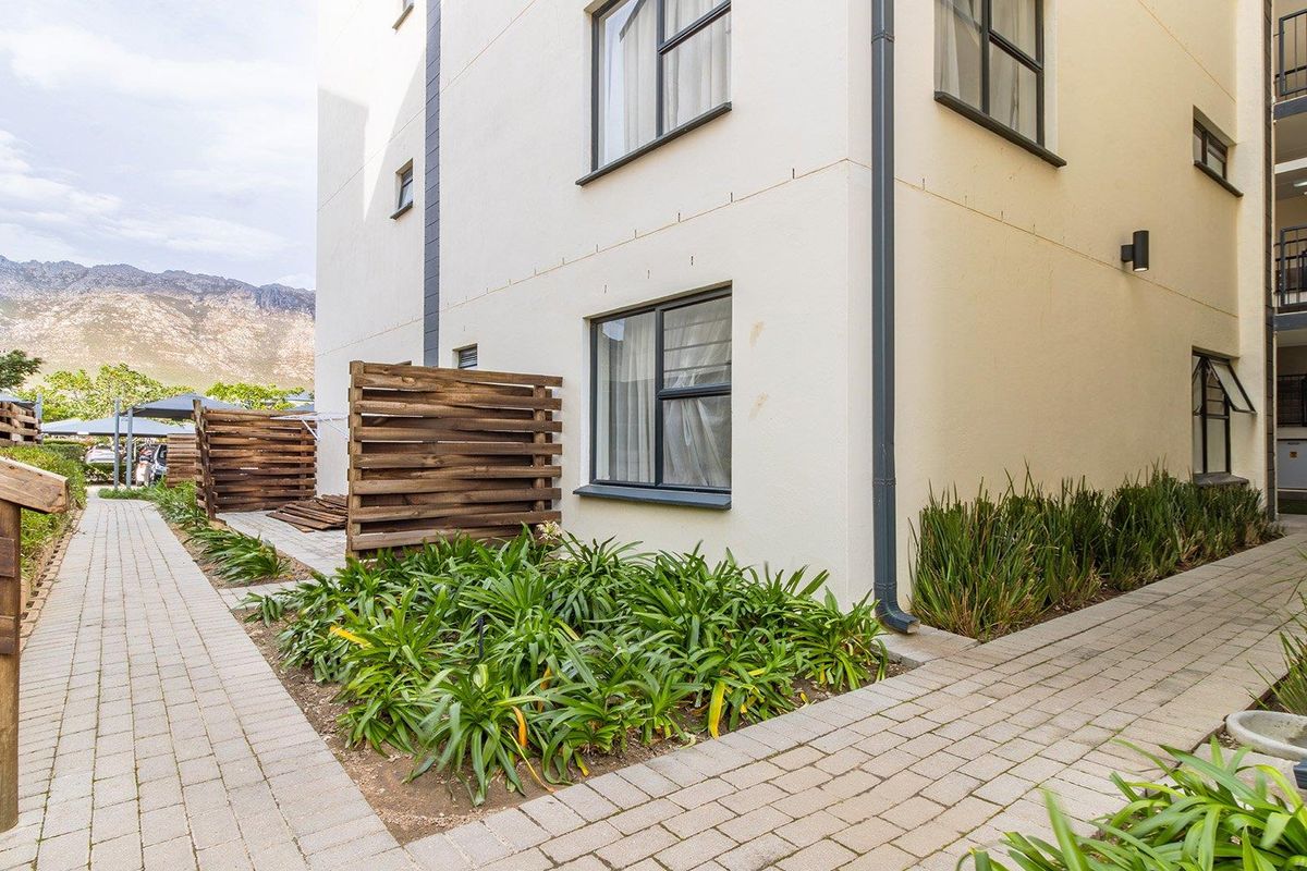 Exterior view of the apartment building featuring paved walkways, landscaped garden beds, and wooden privacy screens with mountain views in the background.