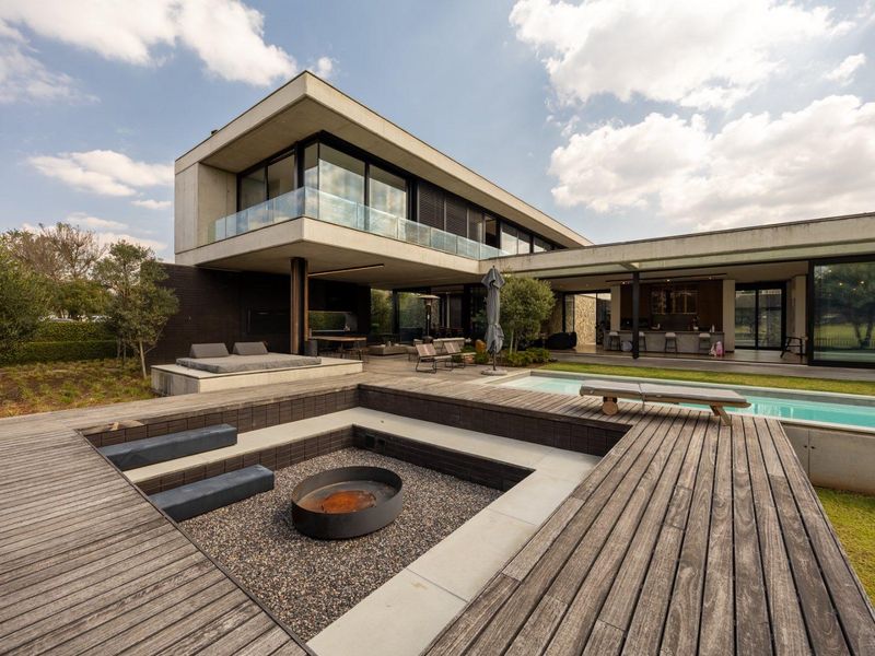 Modern architectural rear exterior featuring a sunken fire pit, extensive timber decking, a swimming pool, and a cantilevered upper level with glass balustrades.