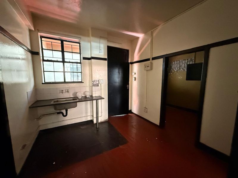 A basic, empty kitchen area featuring a stainless steel sink, tiled splashback, and painted concrete flooring, requiring significant cosmetic updates.